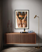 A framed art print featuring a close-up photograph of a person in black lingerie with a gold cross necklace, displayed on a gray wall above a wooden sideboard in a styled room setting.