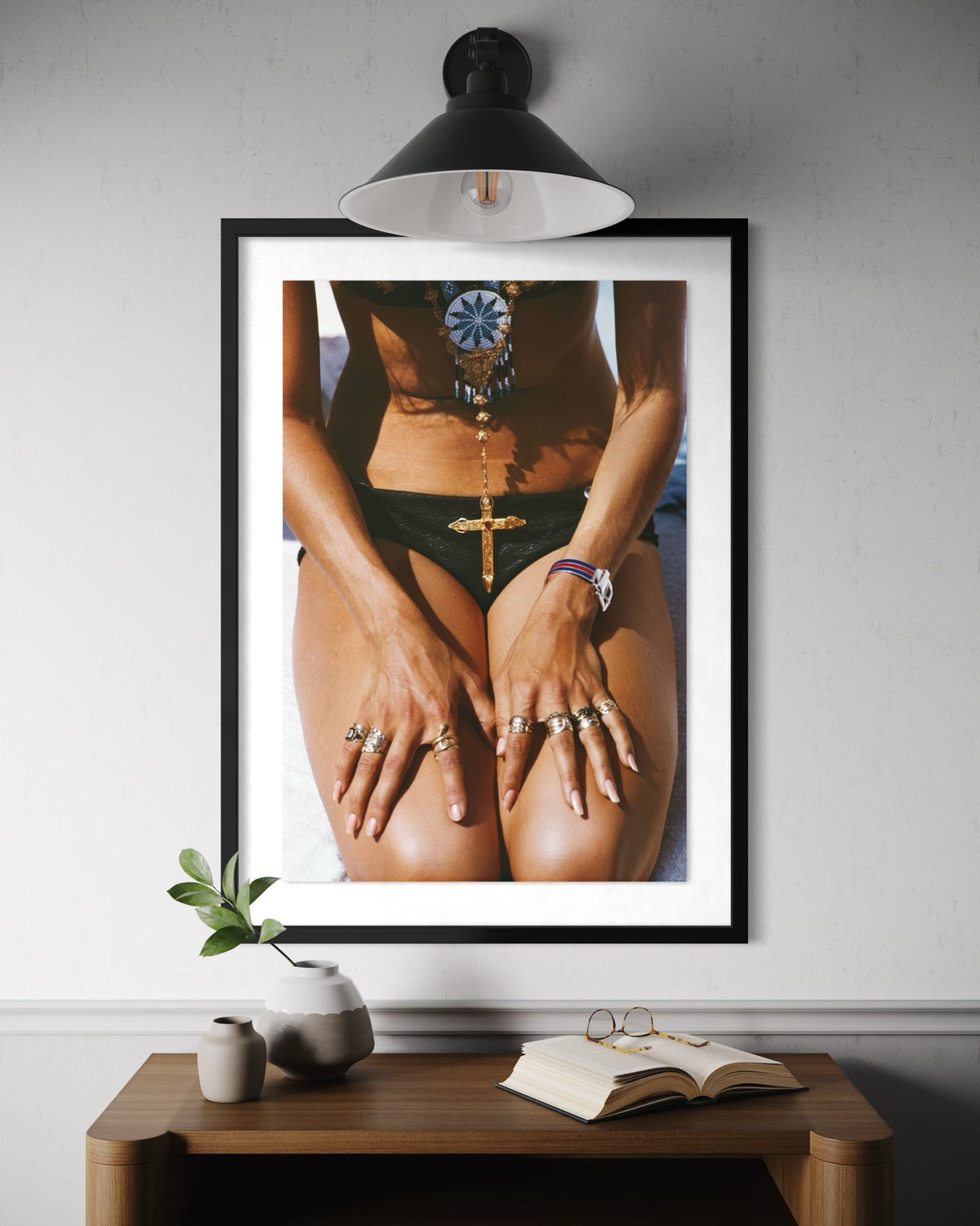 A framed art print featuring a close-up photograph of a person wearing black underwear and multiple rings, hung on a light gray wall above a wooden console table with a lamp, vase, and open book.
