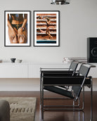 A styled interior room showing two framed art prints hanging on a white wall above a white console. The left print features a close-up of a person's torso in a green bikini, and the right print shows a woman standing on wooden steps wearing a bikini.