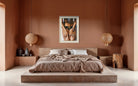A modern bedroom with a low platform bed, brown bedding, and a terracotta-colored wall featuring a framed art print of a person's torso. Two spherical pendant lights hang on either side of the bed.