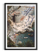 A framed art print depicting a scenic coastal village and beach with a stone tower, rocky cliffs, blue water, and beachgoers, shown against a clean white background.