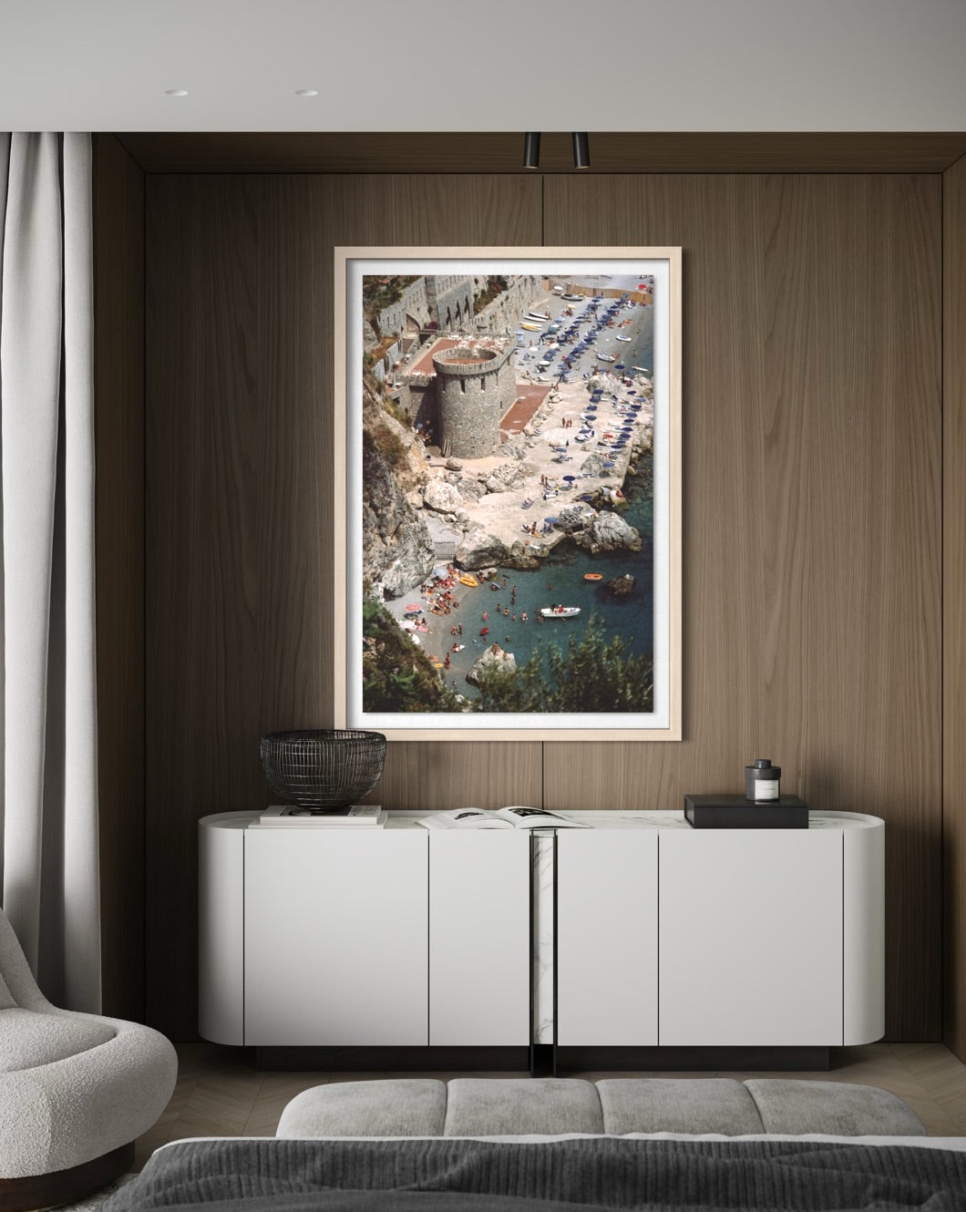 A framed vertical art print depicting a coastal seaside resort with a stone tower, blue pool water, and beach umbrellas, displayed on a dark wood wall above a white sideboard in a modern room.
