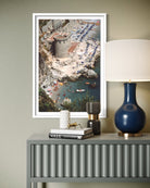 A framed vertical art print depicting an aerial view of a coastal beach and harbor with a stone tower, displayed on a gray console table next to a blue lamp and books.