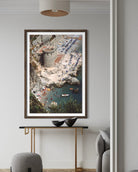 A framed vertical art print depicting a coastal beach and harbor scene with boats, hung on a light gray wall above a console table in a modern living space.