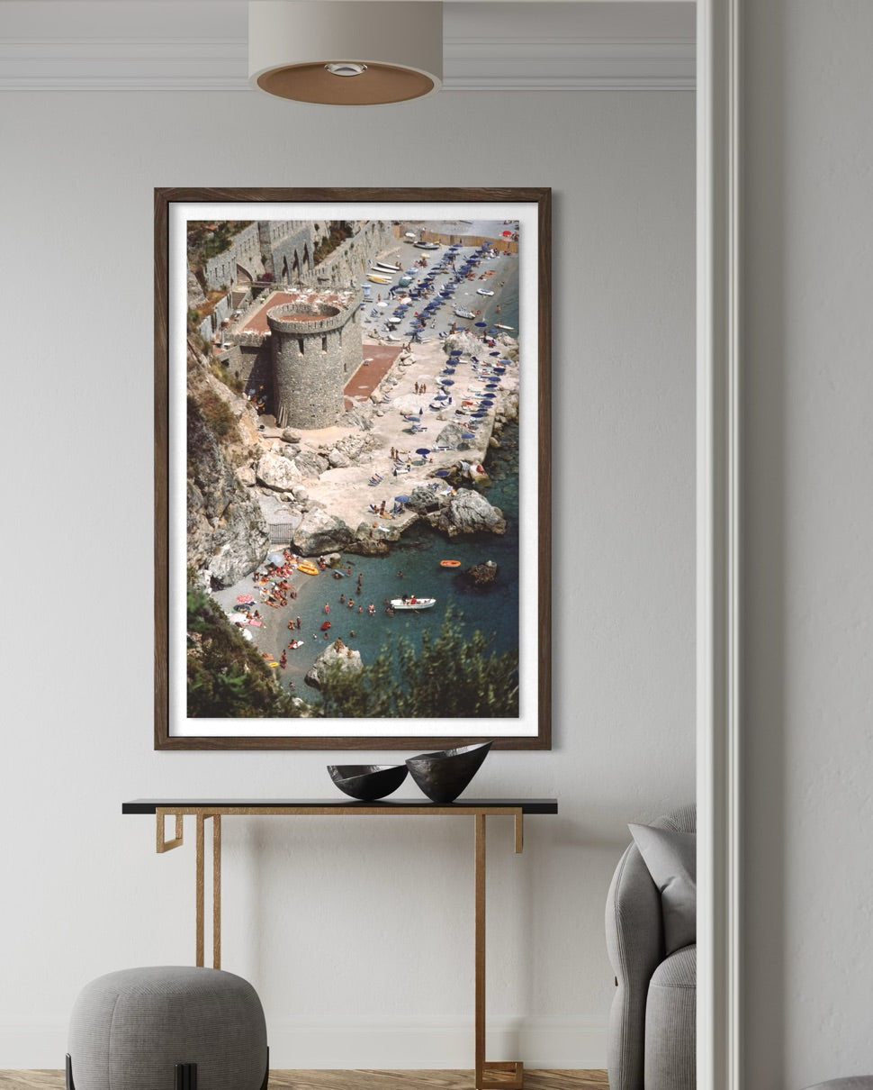 A framed vertical art print depicting a coastal beach and harbor scene with boats, hung on a light gray wall above a console table in a modern living space.