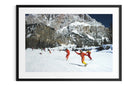 A framed art print featuring a winter mountain scene with several people in bright orange ski suits gliding on a snowy slope under a dark blue sky.