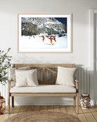 A framed art print depicting skiers in bright orange outfits against a snowy mountain landscape, displayed on a white wall above a wooden bench with neutral-toned cushions in a styled interior setting. The Picturalist Wall Art and Photography for Designed Interiors. Slim Aarons Collection, Skiing. Getty Images Gallery. 