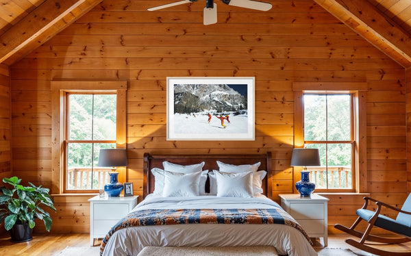 A cozy mountain cabin bedroom with wood-paneled walls, a bed with white linens and a patterned blue-orange throw, two windows, bedside tables with blue lamps, and a framed winter landscape print above the bed. The Picturalist Wall Art and Photography for Designed Interiors. Slim Aarons Collection, Skiing. Getty Images Gallery. 
