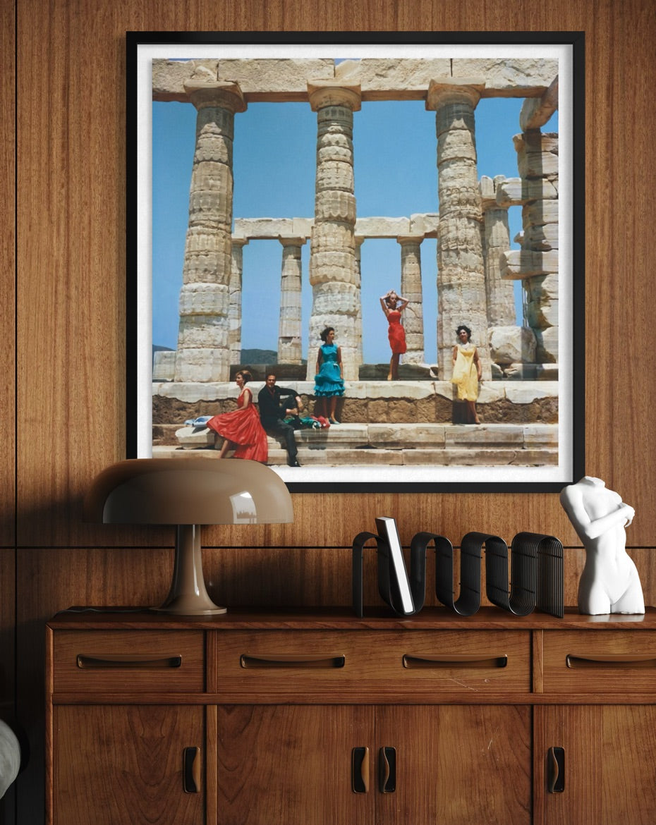 A framed art print depicting ancient stone columns and figures in a temple scene, displayed on a wooden wall above a matching wooden cabinet with a lamp and decorative objects.