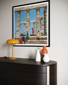 A framed art print depicting the ancient Greek Temple of Poseidon at Sounia, Greece, styled on a light gray wall above a dark wood console table with a yellow lamp and ceramic vases.