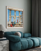 A framed art print depicting ancient stone columns and figures in colorful garments, displayed on a sage green wall above a modern teal sofa in a styled living room.