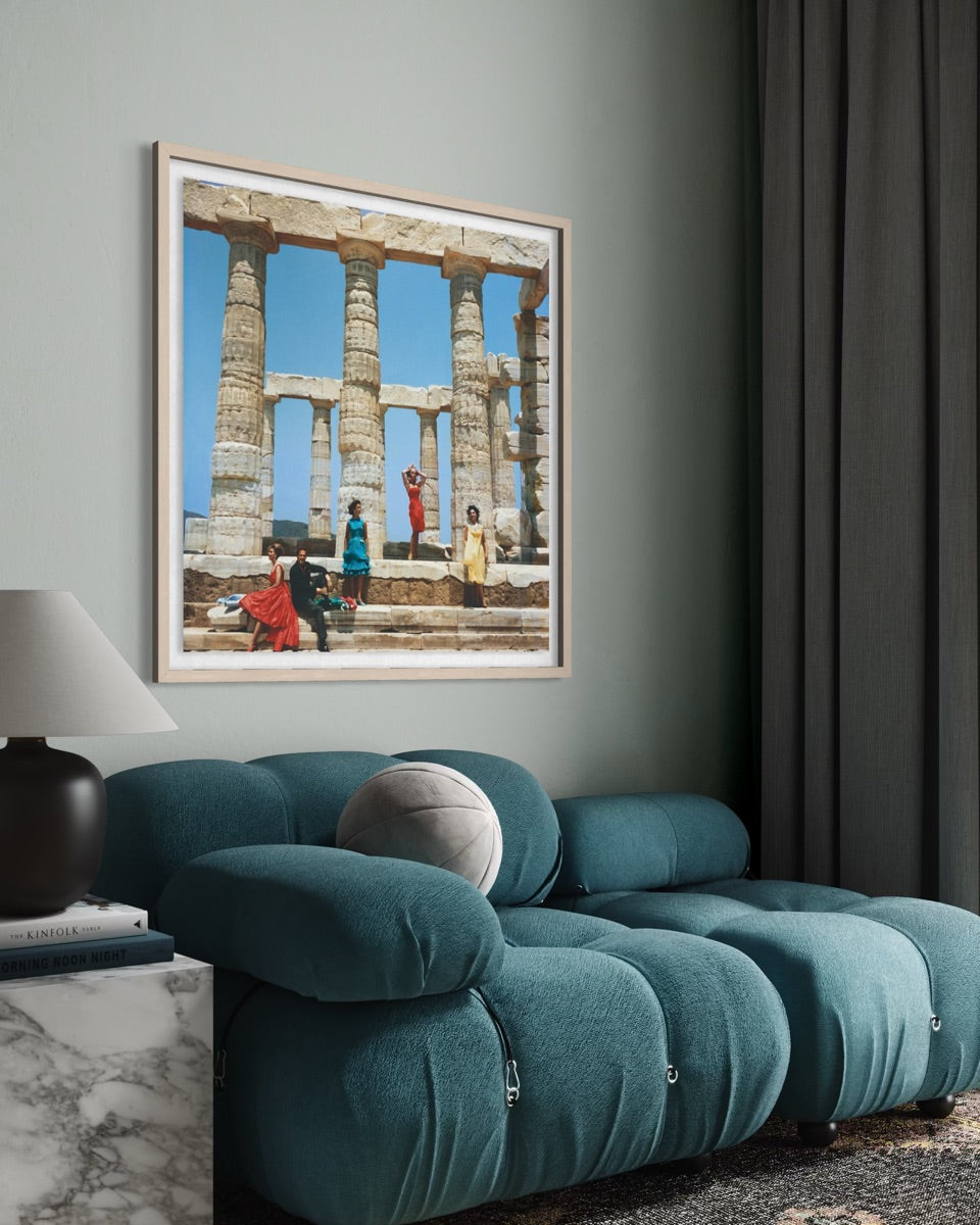 A framed art print depicting ancient stone columns and figures in colorful garments, displayed on a sage green wall above a modern teal sofa in a styled living room.