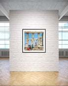 A framed art print depicting ancient stone columns and figures under a blue sky, displayed on a white brick wall in a modern room with wood floors.