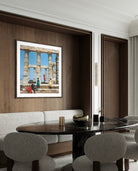 A framed art print depicting ancient stone columns and figures under a blue sky, displayed on a dark wood-paneled wall above a dining table with gray upholstered chairs.