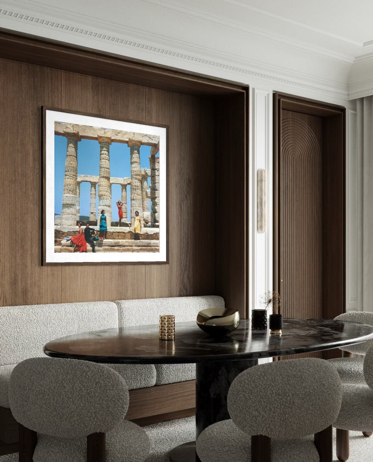 A framed art print depicting ancient stone columns and figures under a blue sky, displayed on a dark wood-paneled wall above a dining table with gray upholstered chairs.
