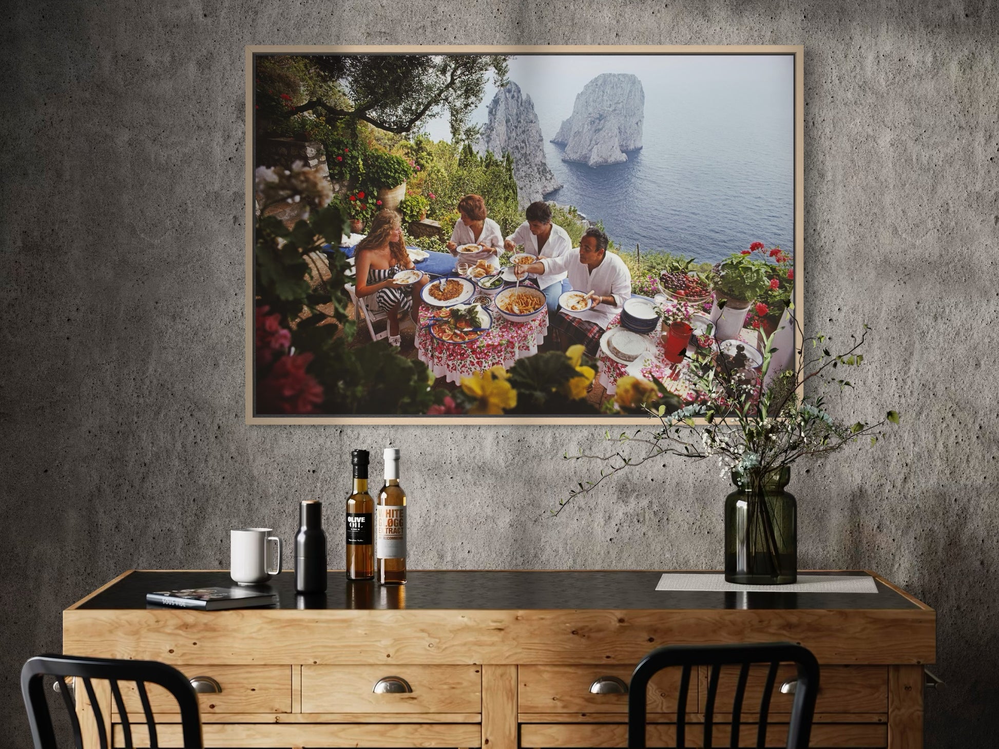 Framed artwork of a group dining outdoors with a scenic background, displayed above a wooden desk.
