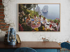 Framed artwork of a group dining outdoors with ocean view on a textured wall.