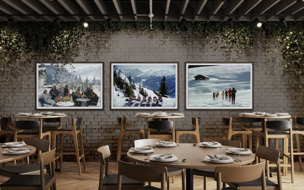 A restaurant interior with three framed winter sports paintings hanging on a brick wall above tables and chairs.
