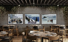 A restaurant interior with three framed winter sports paintings hanging on a brick wall above tables and chairs.
