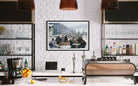 A framed art print depicting a group of people dining outdoors in a snowy mountain setting, displayed on a white brick wall above a modern kitchen counter with coffee equipment and bar shelves.