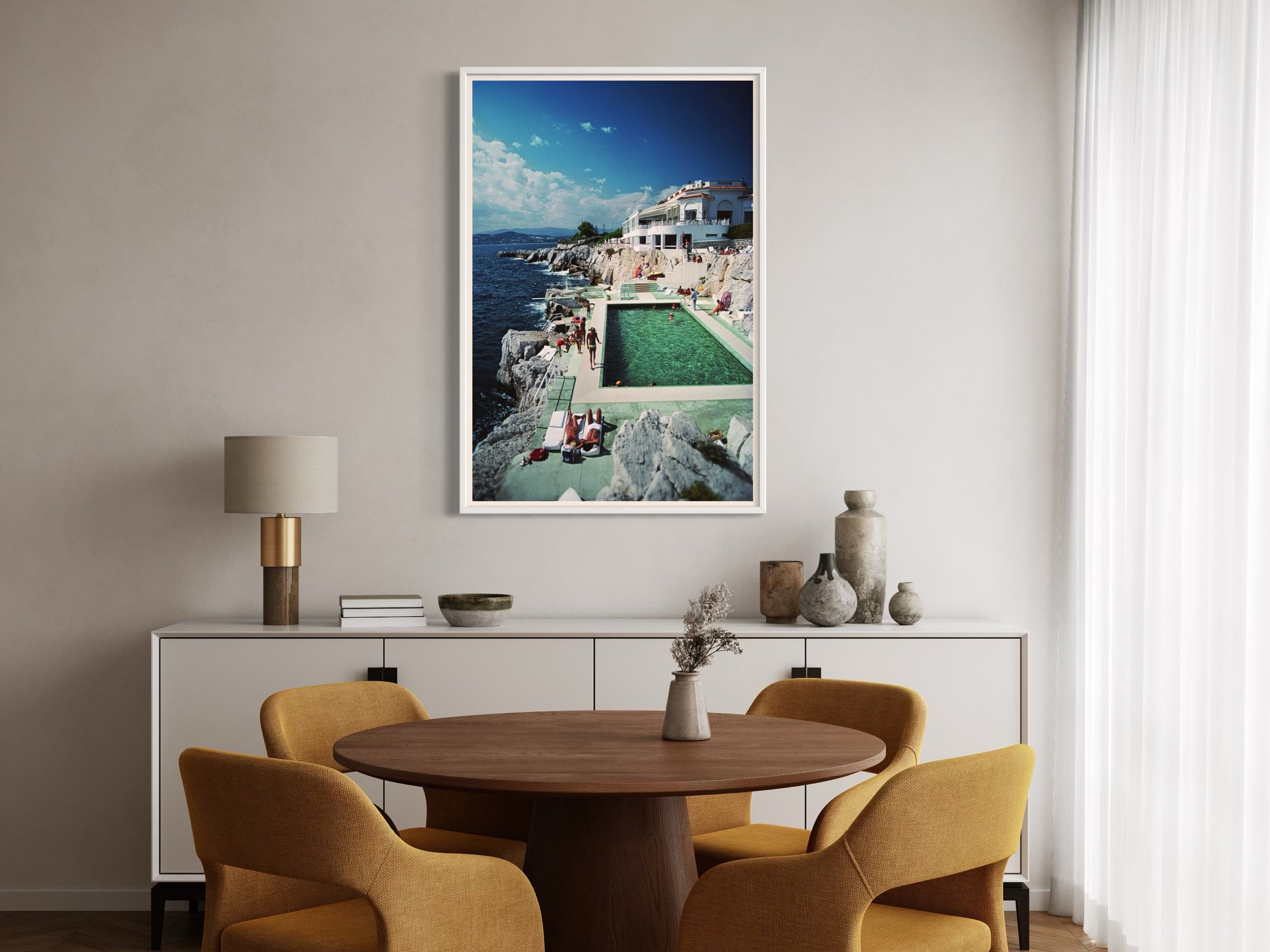 Dining room with a round wooden table and yellow chairs, featuring a framed picture of a poolside scene on the wall.