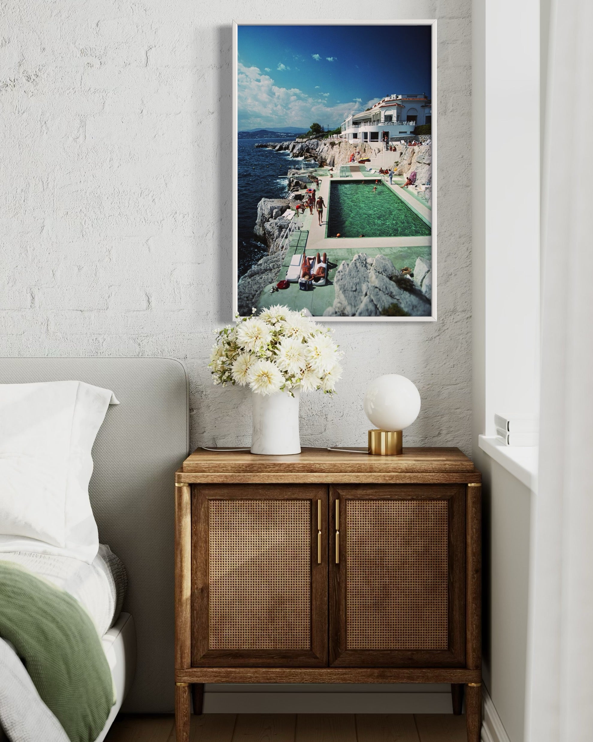 Wooden side table with a vase of flowers and a lamp, featuring a large framed picture of a pool scene on a white wall.