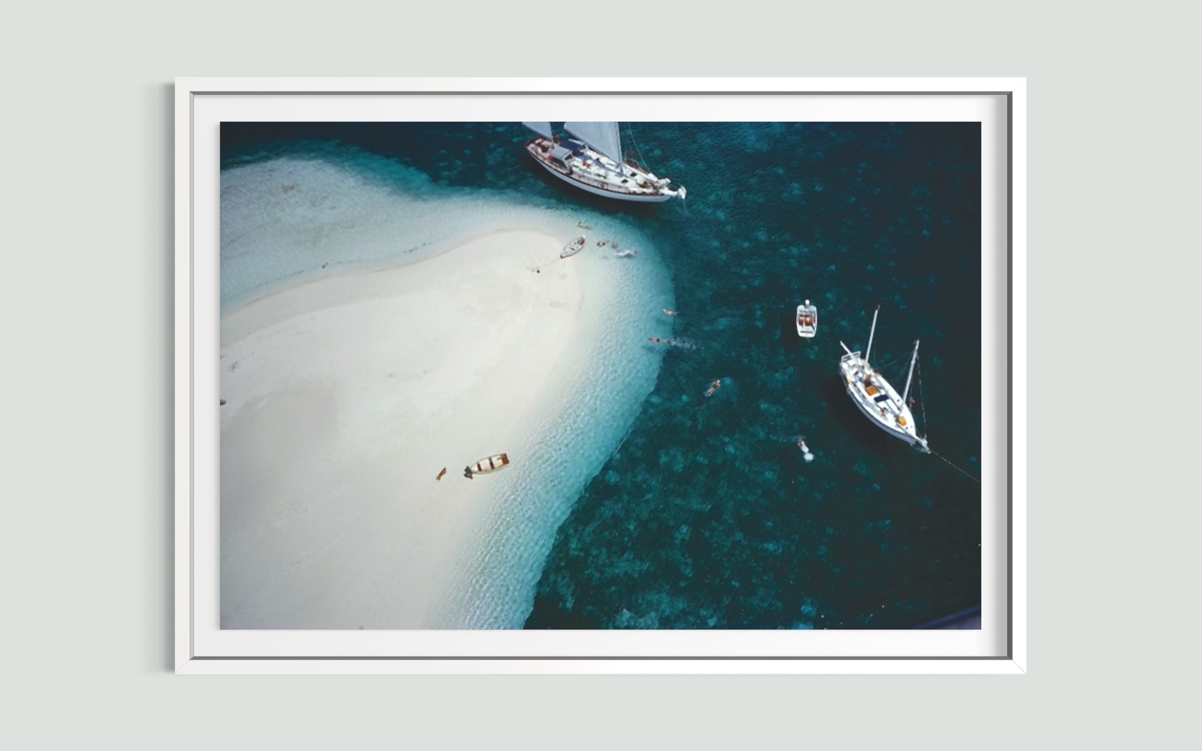 A framed aerial photograph of a white sandbar winding through deep blue and teal water, with several sailboats anchored nearby, displayed on a light gray wall.