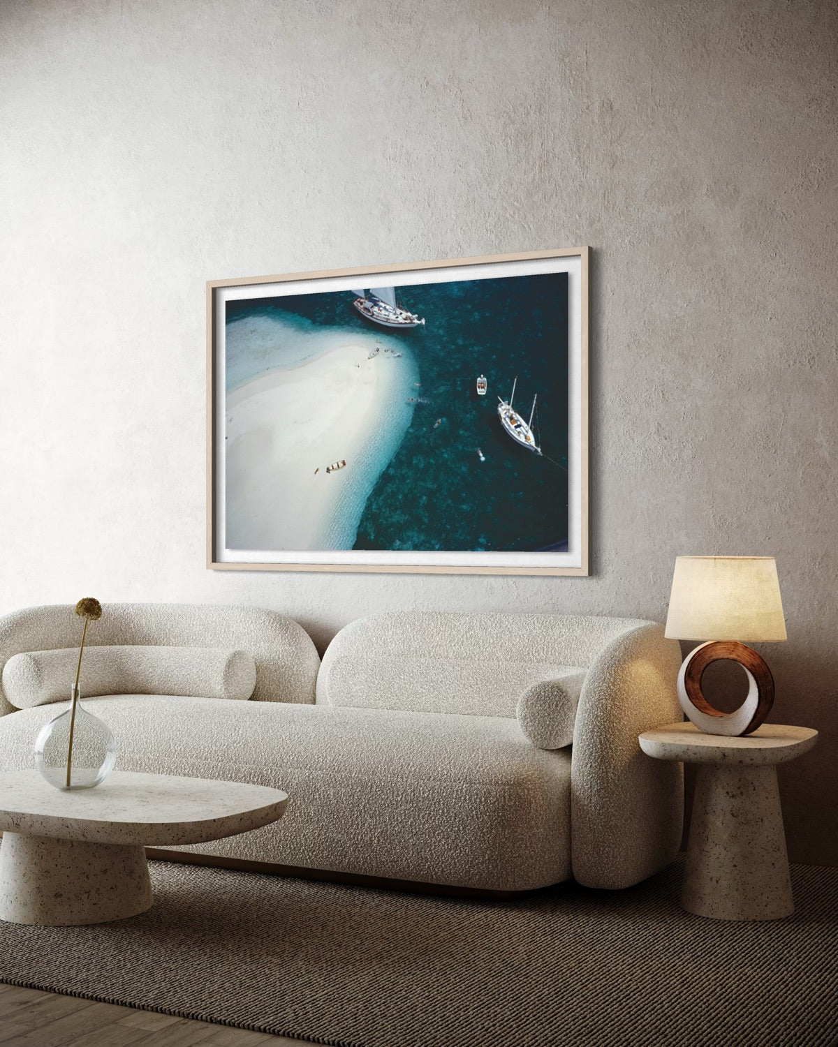 A framed aerial photograph of a beach and boats hanging on a textured beige wall above a modern curved sofa in a styled living room. The Picturalist Wall Art and Photography for Designed Interiors. 