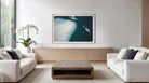 A framed square art print depicting an aerial view of a white sand beach and deep blue water, displayed on a light wood console in a modern, minimalist living room with white sofas and a wooden coffee table. The Picturalist Wall Art and Photography for Designed Interiors.