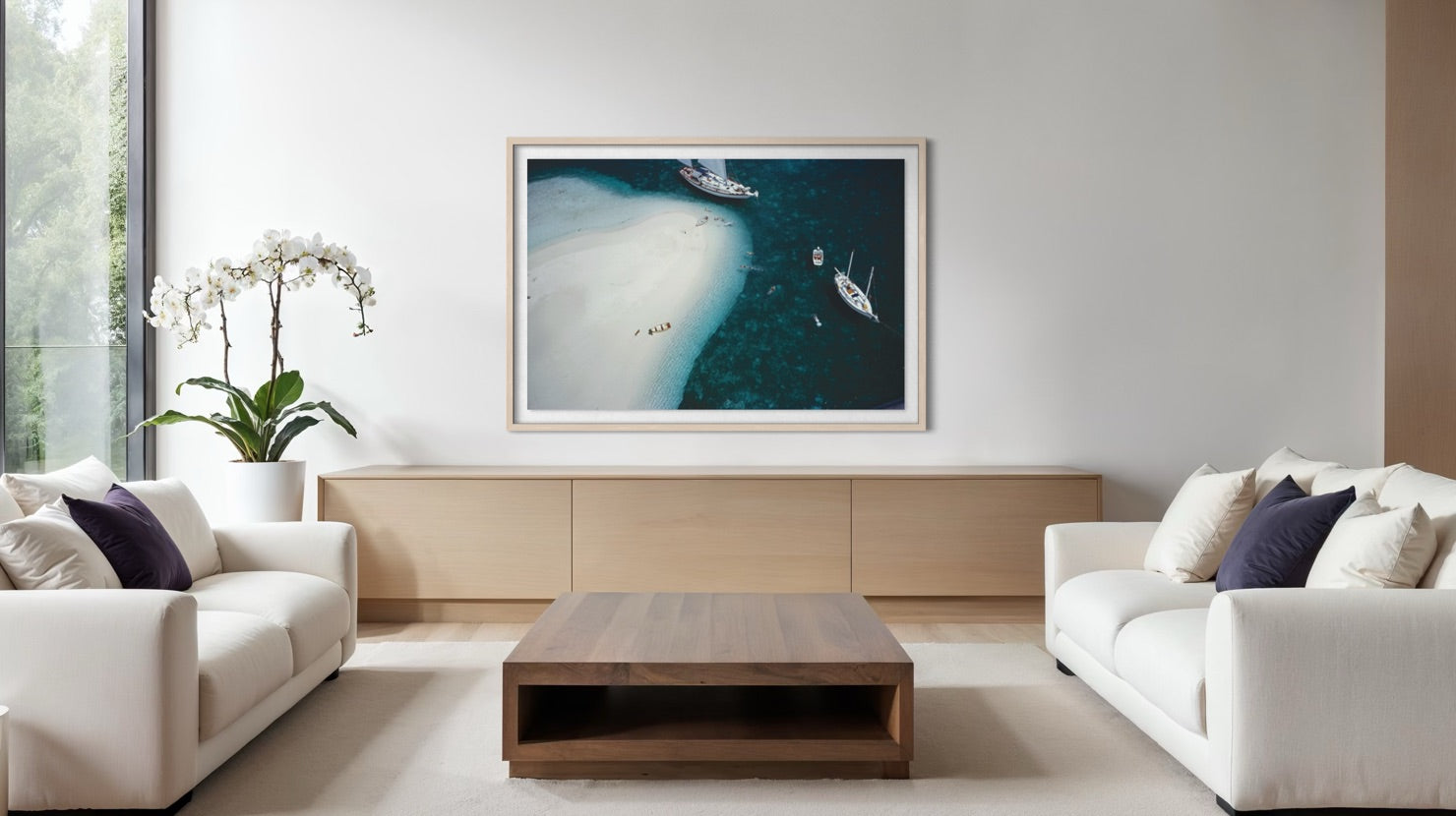 A framed square art print depicting an aerial view of a white sand beach and deep blue water, displayed on a light wood console in a modern, minimalist living room with white sofas and a wooden coffee table. The Picturalist Wall Art and Photography for Designed Interiors.