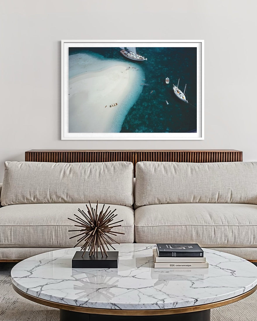 A framed aerial photograph of a white sand beach and sailboats in deep blue water, displayed on a light gray wall above a beige sofa in a modern living room setting. The Picturalist Wall Art and Photography for Designed Interiors. 