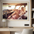 A framed art print depicting a blonde woman in a vintage car interior, displayed on a light-colored wall above a wooden desk with a lamp and chair.