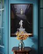 Framed artwork of a classical statue on a blue wall with a vase of flowers below.