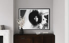 Framed black and white portrait of a person with an afro on a wall above a wooden cabinet.