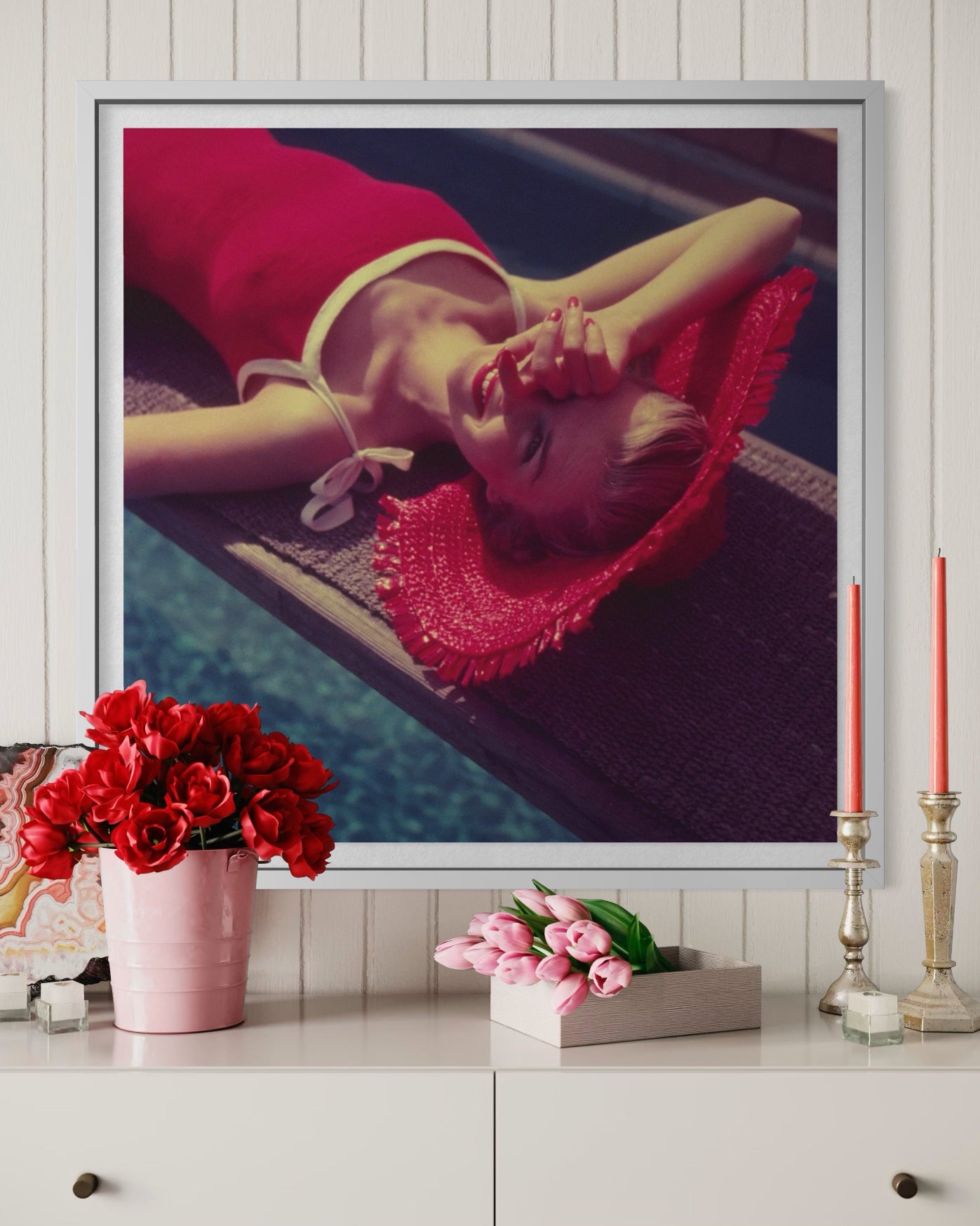 Framed artwork of a woman in a red dress and hat on a wall above a decorative table with flowers and candles.