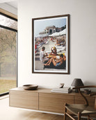 A framed vertical art print depicting a sunny beach scene with people lounging under white umbrellas, displayed on a white wall above a light wood console table in a modern interior setting.