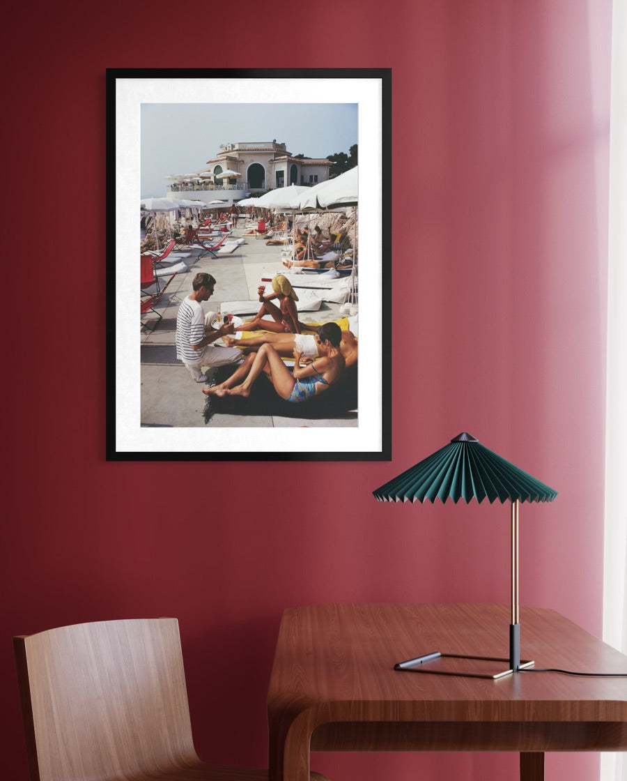 A framed art print depicting a vintage beach scene with people lounging under umbrellas, hanging on a deep red wall above a wooden table with a green pleated lamp.