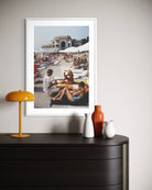 A framed art print depicting a sunny beach scene with people lounging under umbrellas, displayed on a light gray wall above a dark wood console table with decorative vases and a yellow lamp.