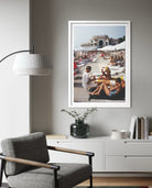 A framed vertical art print depicting a crowded beach scene with people lounging under umbrellas, displayed on a gray wall above a white console table in a modern living room setting.