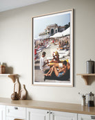 A framed vertical art print depicting a sunny beach scene with people lounging by the pool, displayed on a beige wall above a kitchen counter with decorative items.