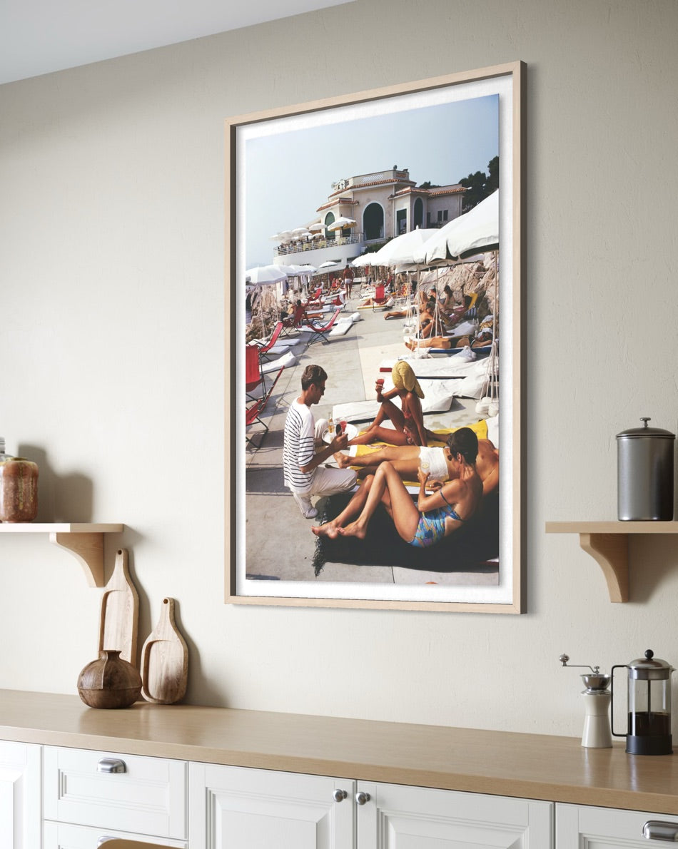 A framed vertical art print depicting a sunny beach scene with people lounging by the pool, displayed on a beige wall above a kitchen counter with decorative items.