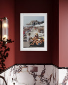 A framed art print depicting a beach scene with people lounging under umbrellas, mounted on a dark red wall powder-room above a marble-patterned surface.