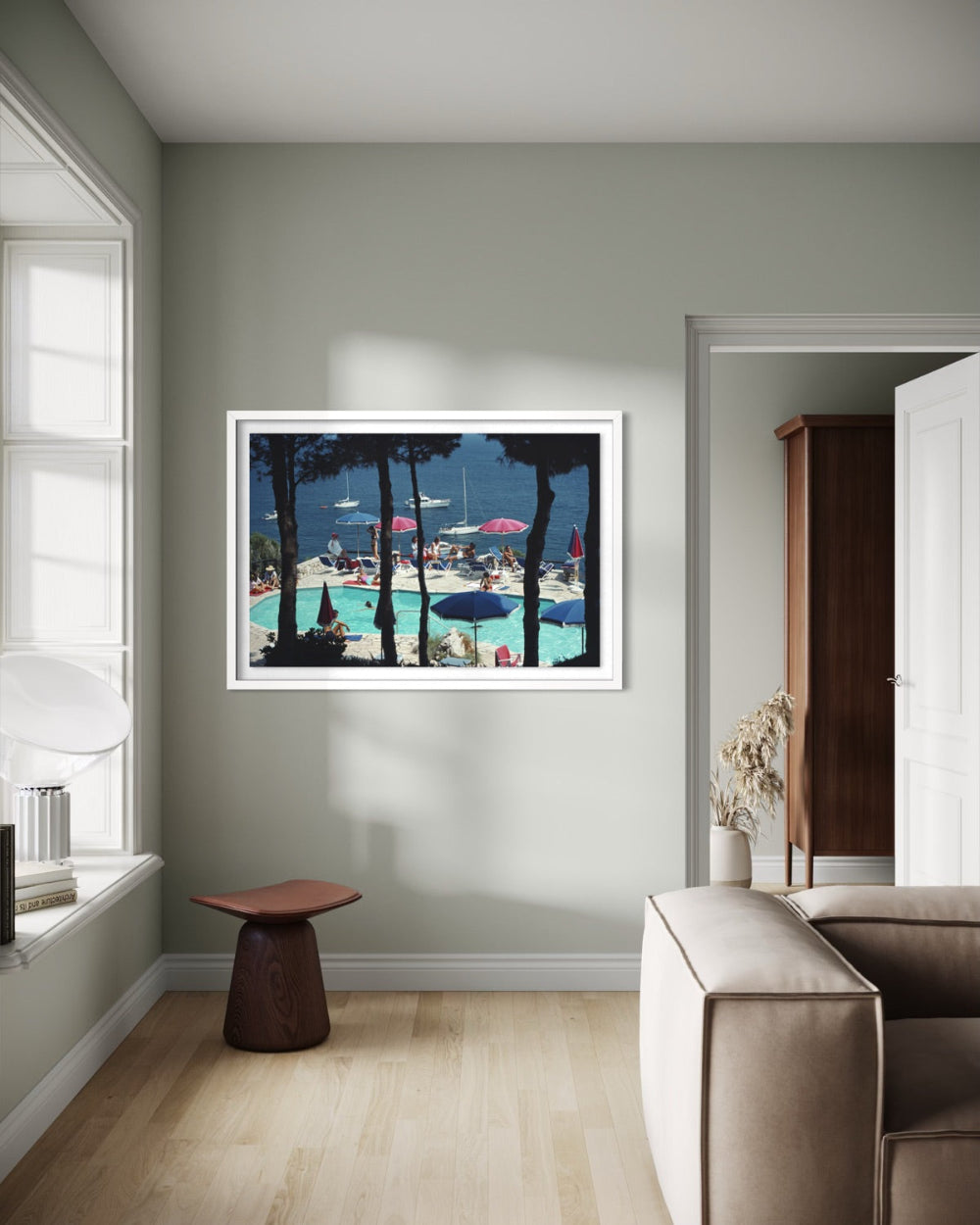A framed art print depicting a vibrant outdoor swimming pool scene with palm trees and beach umbrellas, displayed on a light sage green wall in a modern living room setting.