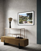 A framed art print depicting a scenic road through lush green hills under a blue sky, displayed on a textured gray wall in a modern living room setting above a gold-framed console table and a tan upholstered bench.