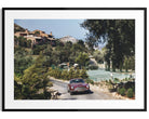 A framed photographic print of a classic red sports car driving down a winding road near tennis courts and hillside villas, presented with a white mat and a black frame.