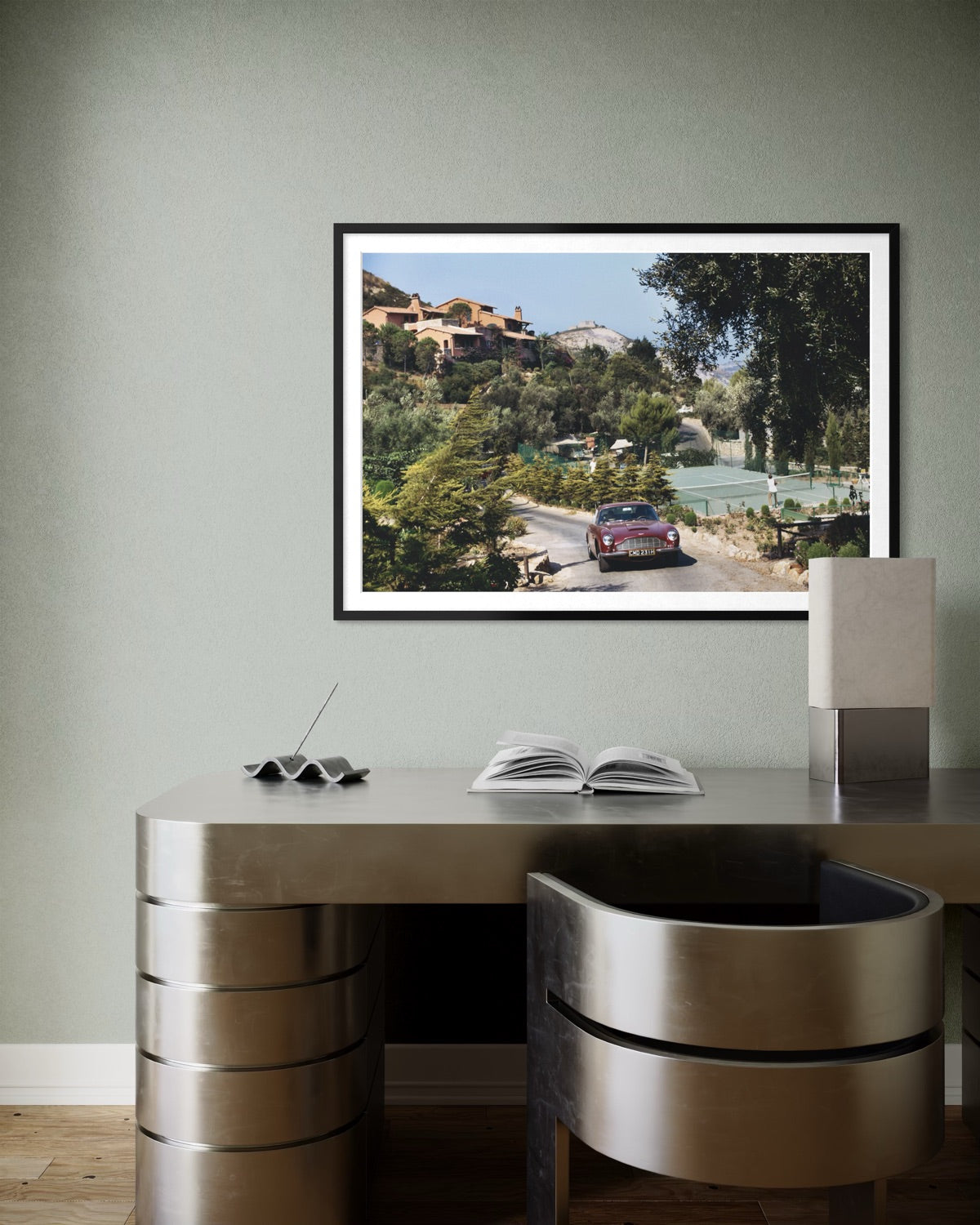 A framed art print depicting a vintage red sports car on a scenic road surrounded by lush green hills and buildings, displayed on a light sage green wall above a modern metallic desk and chair.