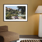A framed art print depicting a scenic Italian hillside road with a classic car, displayed on a warm beige wall in a modern living room setting next to a brown leather sofa and a white floor lamp.