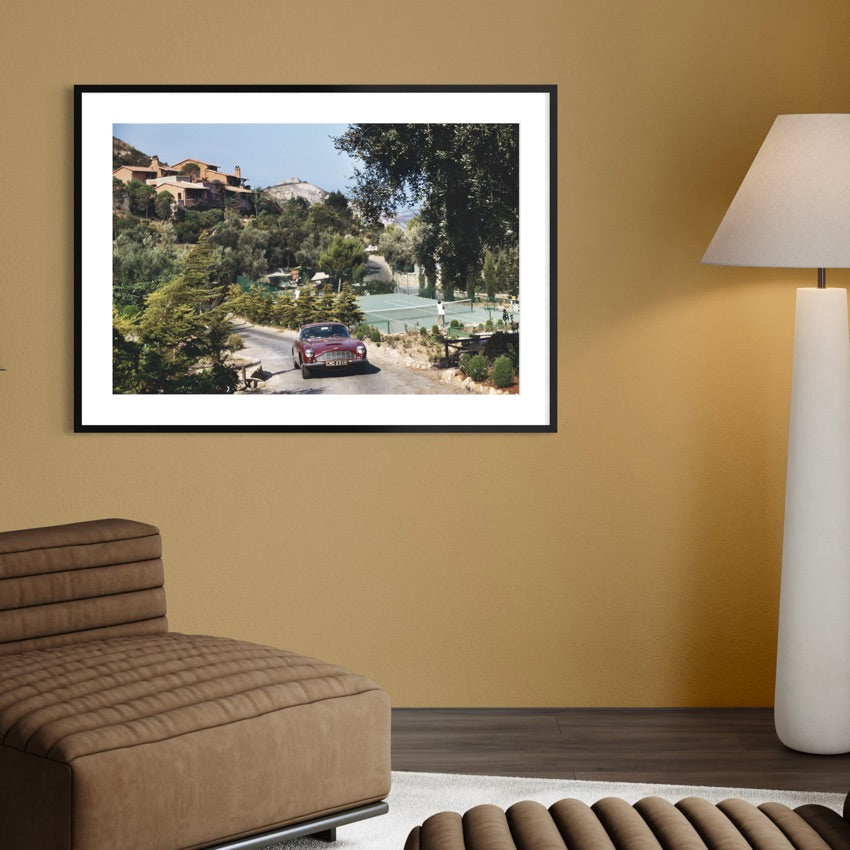 A framed art print depicting a scenic Italian hillside road with a classic car, displayed on a warm beige wall in a modern living room setting next to a brown leather sofa and a white floor lamp.