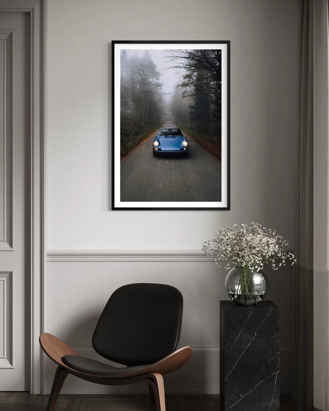 A framed vertical art print depicting a blue classic car driving down a misty forest road, displayed on a gray wall in a modern living room setting above a black chair and next to a vase of flowers.