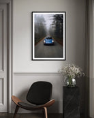 A framed vertical art print depicting a blue classic car driving down a misty forest road, displayed on a gray wall in a modern living room setting above a black chair and next to a vase of flowers.